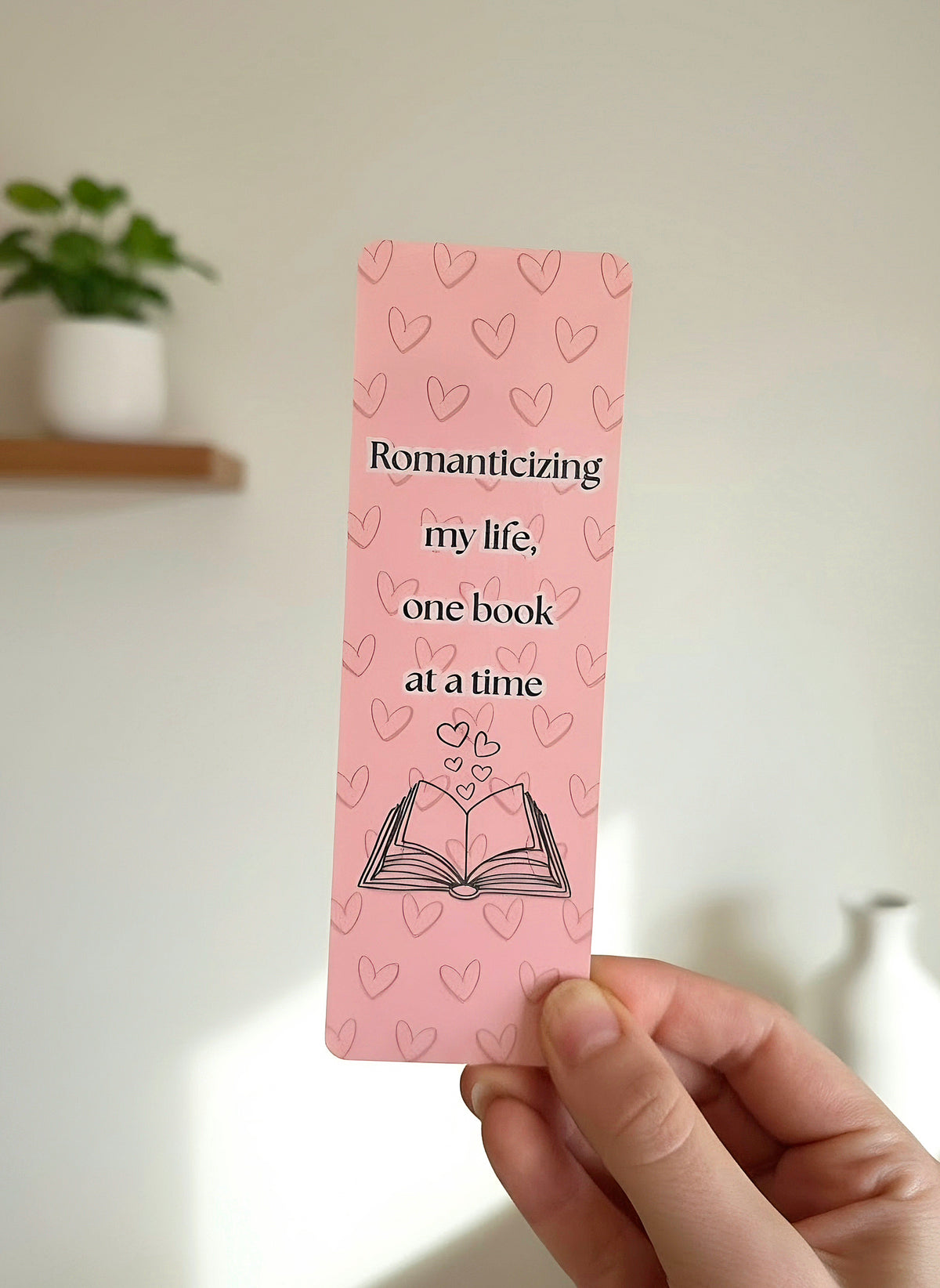 A hand holding a pink bookmark with a heart pattern and the words "Romanticizing my life, one book at a time" on the front. Includes an open book and hearts coming out of the book. 