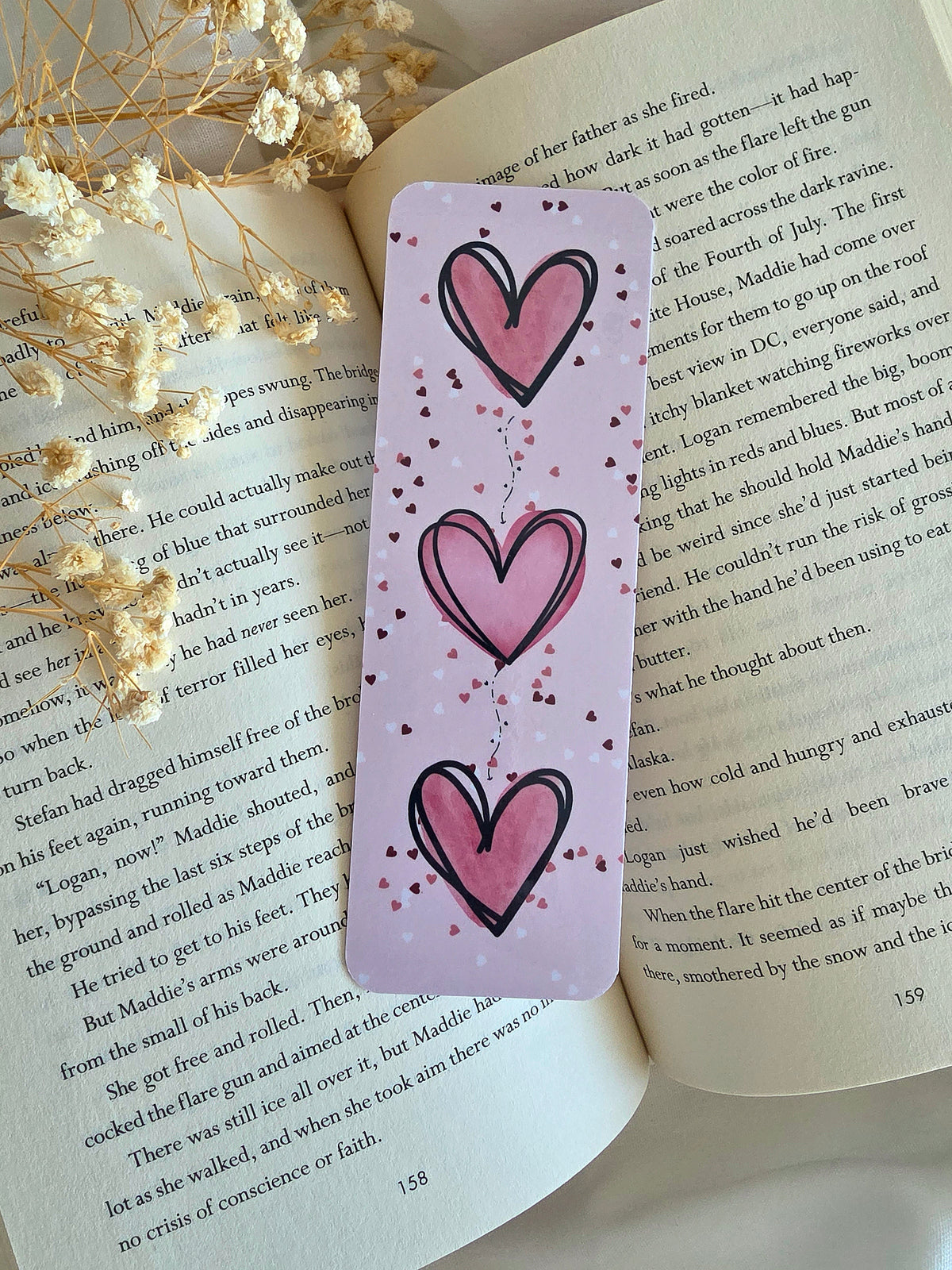A pink bookmark with three big pink hearts and white, pink, and dark red heart confetti print. The bookmark is laying on an open book with some baby's breath flowers on the left.
