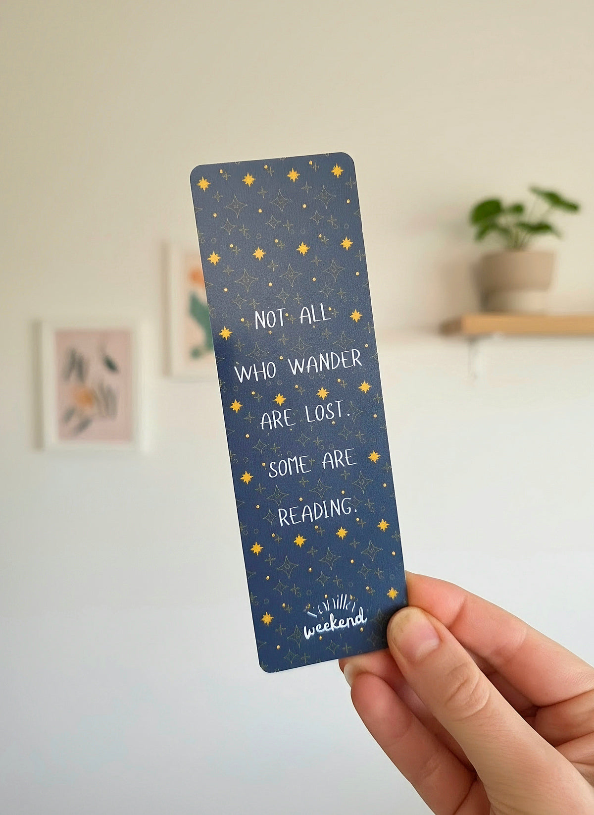 A hand holding a bookmark with a dark blue background, gold star pattern, and with a text overlay saying "Not all who wander are lost. Some are reading." In the background of the photo there are wall photos and a potted plant on a shelf.
