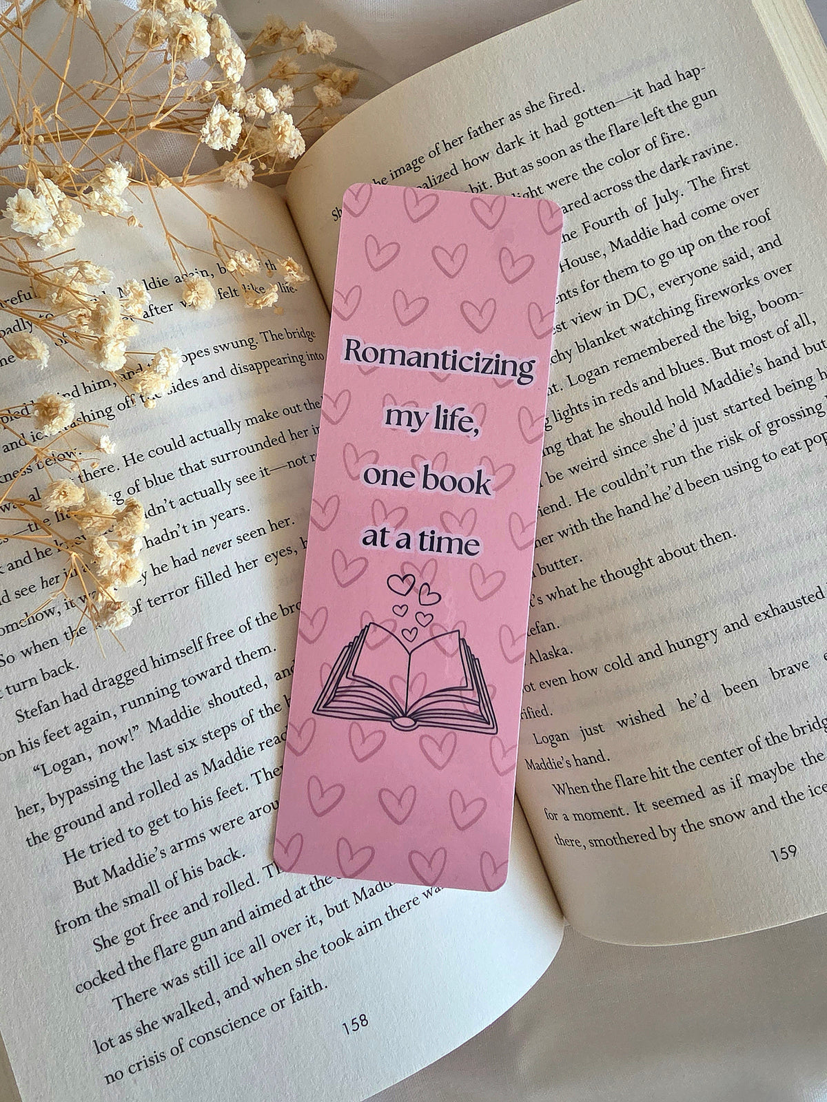 A pink bookmark with a heart pattern and the words "Romanticizing my life, one book at a time" on the front. Includes an open book and hearts coming out of the book. The bookmark is laying on an open book with some baby's breath flowers on the left.