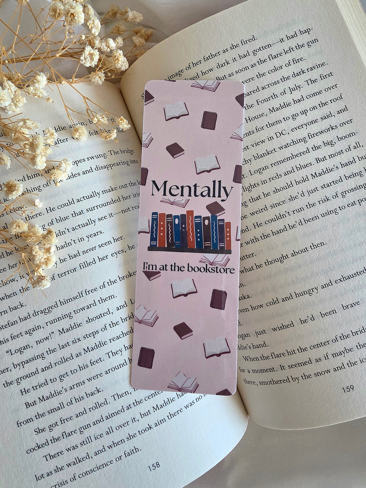 A cream colored bookmark with books in the background. An overlay of "Mentally I'm at the bookstore" is written on the front with a shelf of blue, orange, and purple books. The bookmark is laying on an open book with some baby's breath flowers on the left.