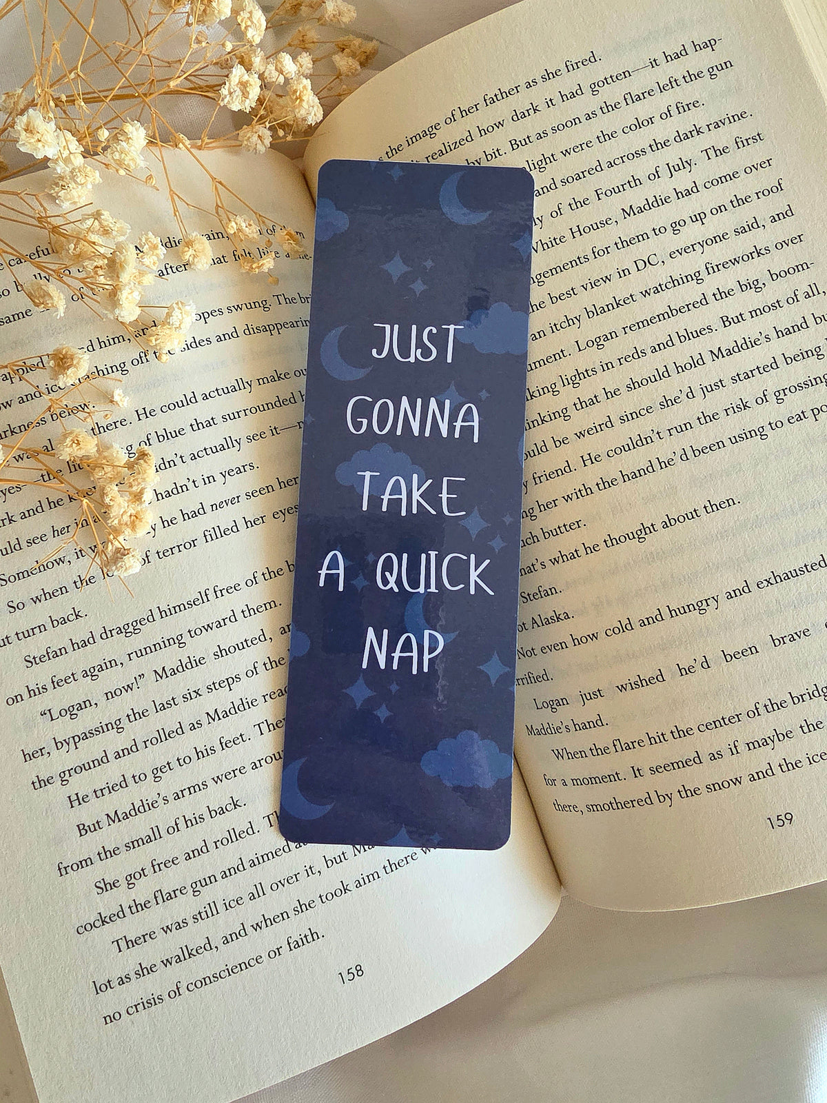 A dark navy blue bookmark with a pattern of the moon, stars, and clouds in the background. An overlay of "Just gonna take a quick nap" is written on the bookmark in white. The bookmark is laying on an open book with some baby's breath flowers on the left.