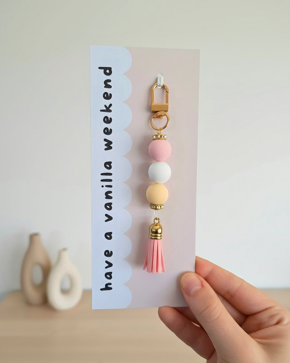 A hand holding a card featuring a keychain with a pink bead, white bead, and yellow bead with gold accents and a pink tassel. The card has the text 'have a vanilla weekend' on a light background.