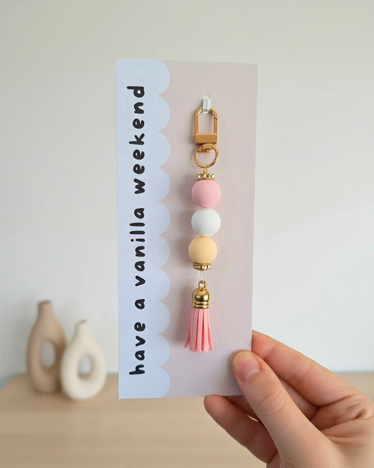 A hand holding a card featuring a keychain with a pink bead, white bead, and yellow bead with gold accents and a pink tassel. The card has the text 'have a vanilla weekend' on a light background.