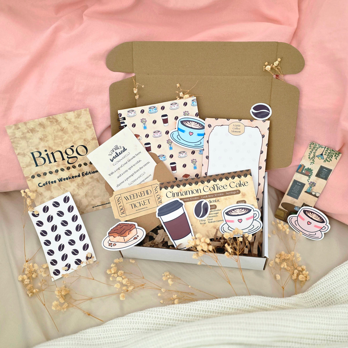 A coffee-themed stationery gift set arranged in an open kraft cardboard box with crinkle paper filler, placed on pink fabric with dried flowers around it. The set includes a spiral notebook, notepad, coffee weekend bingo card with coffee bean stickers, recipe card, double sided bookmark, sticker pack with 7 stickers, and an envelope to open with journal prompts and a collectible weekend ticket. 