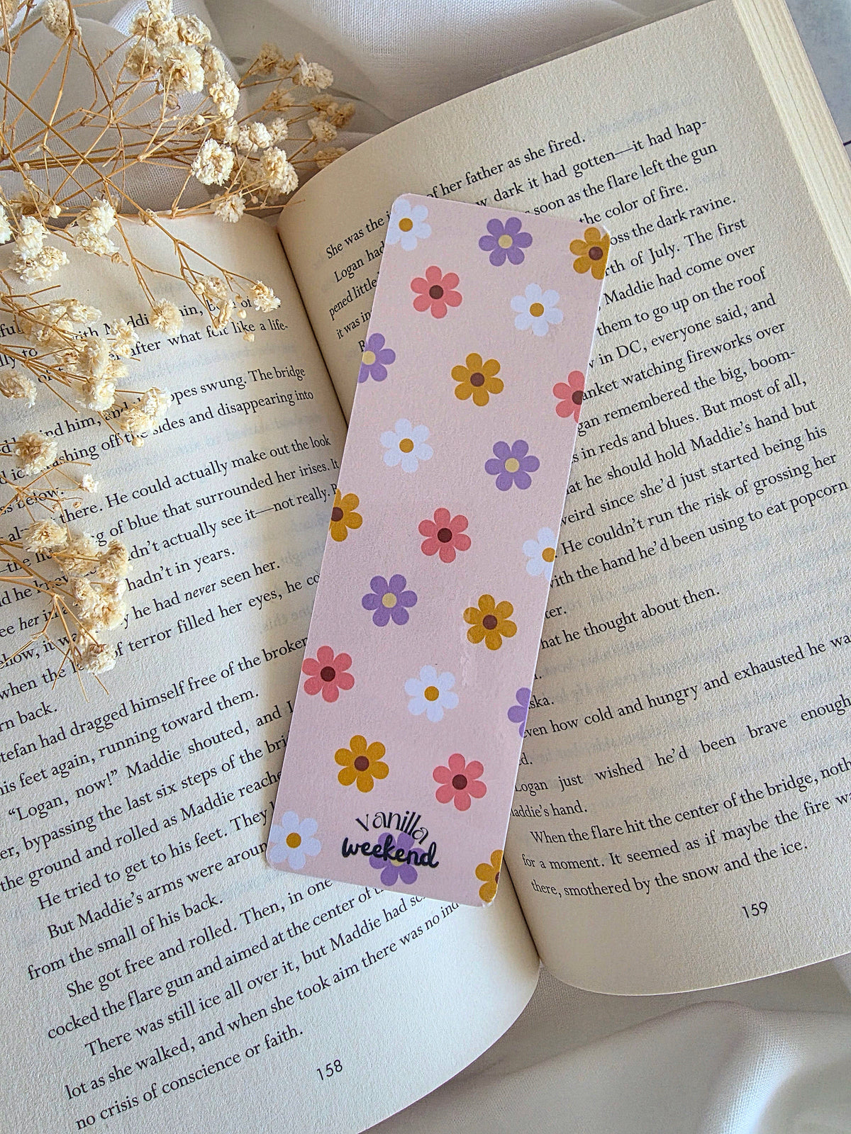 A light pink / cream bookmark with simple flower pattern design with yellow, pink, white, and purple daisies. The bottom says Vanilla Weekend. The bookmark is laying on an open book with baby's breath flowers on the left.