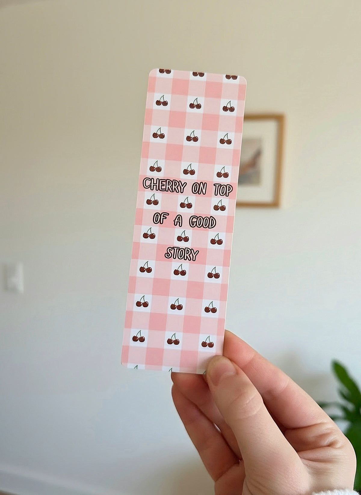 A hand holding a bookmark with a pink and white gingham pattern with mini cherries in the white boxes. An overlay of "Cherry on Top of a Good Story" is written in the middle. 