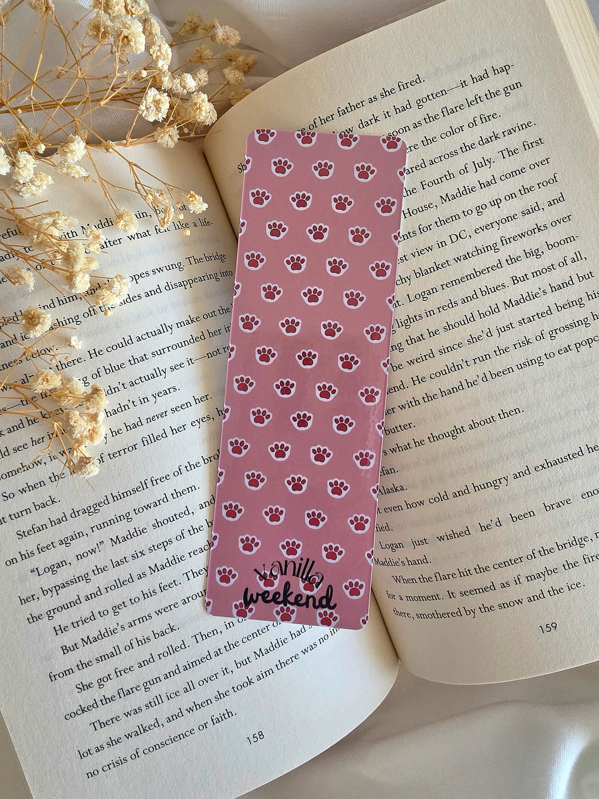A bookmark with a pink background and light pink kitty paw pattern. Bookmark is laying on an open book with some baby's breath flowers on the left.